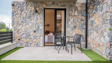 Pared exterior de piedra, una puerta de vidrio que da a una habitación con cama y mesa, y un pequeño patio con sillas y césped artificial.