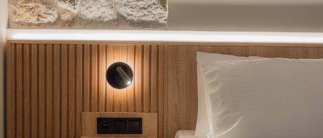 A bedroom with a wooden headboard, a bedside table with a lamp, a stone wall, and a bed with white bedding.