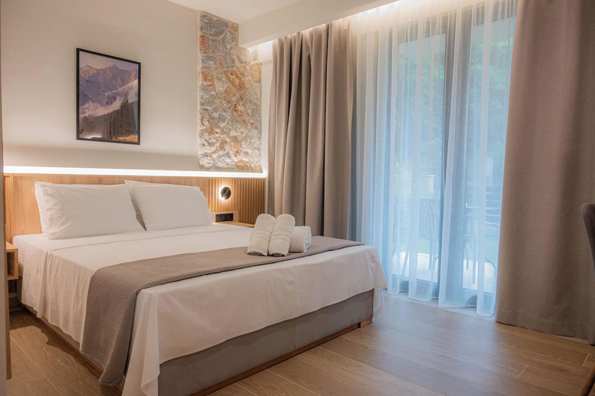 A hotel room with a bed, a nightstand, a wall-mounted lamp, a framed picture, and a large window with curtains.