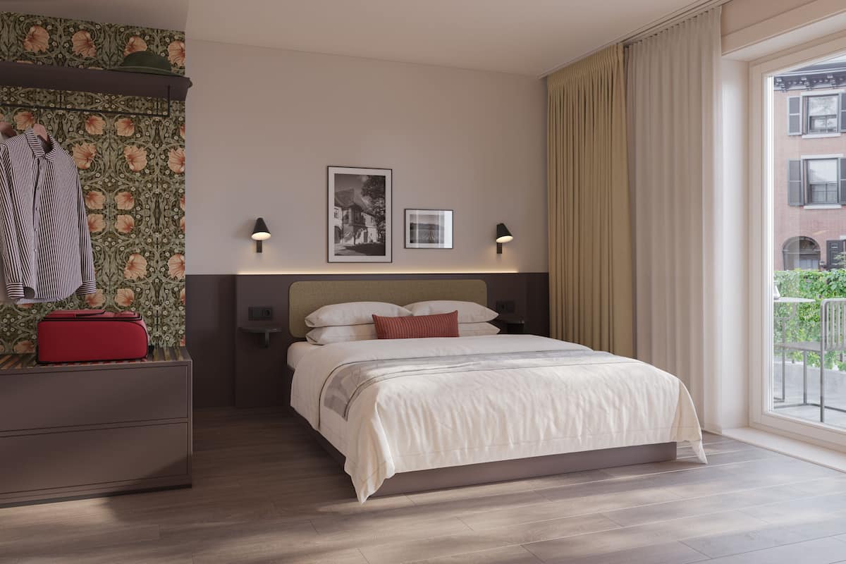A hotel room with a bed, a wardrobe, a red suitcase, two wall-mounted lamps, and a window with a view of buildings.