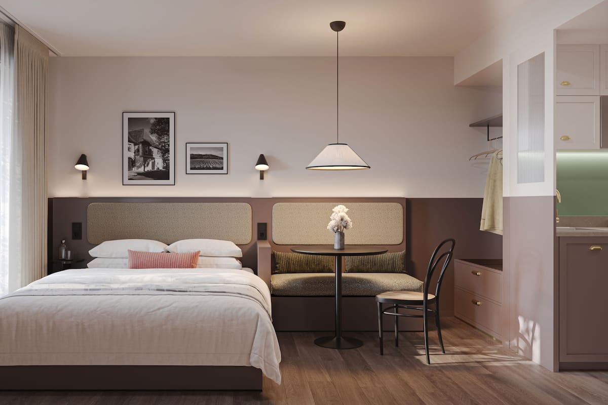A hotel room with a bed, a small table with a chair, a sofa, a wall-mounted lamp, and a framed picture.