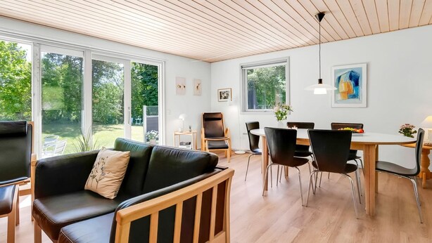 Uma sala de estar moderna com teto de madeira, grandes janelas, um sofá de couro preto e uma área de jantar com mesa e cadeiras de madeira.