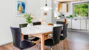 Uma cozinha moderna com armários brancos, uma área de jantar com mesa redonda e cadeiras pretas, e uma janela grande com vista para o verde.