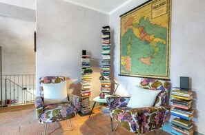Ein Zimmer mit zwei blumigen Sesseln, einem hohen Bücherstapel, einer Karte an der Wand und einer Treppe im Hintergrund.