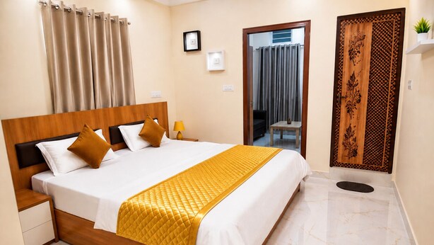 A hotel room with a large bed, a wooden headboard, a bedside table, a nightstand, a mirror, a door with intricate woodwork, and a small potted plant. - Ferro Livingz (Bengaluru)