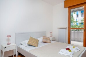 Ein Schlafzimmer mit einem Bett, zwei Kissen, einem Nachttisch mit Lampe und Blick ins Freie durch eine Tür.