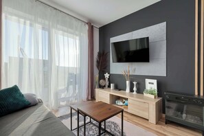 Ein modernes Wohnzimmer mit einer Couch, einem Couchtisch, einem an der Wand montierten Fernseher, einem Kamin und einem Fenster mit durchsichtigen Vorhängen.