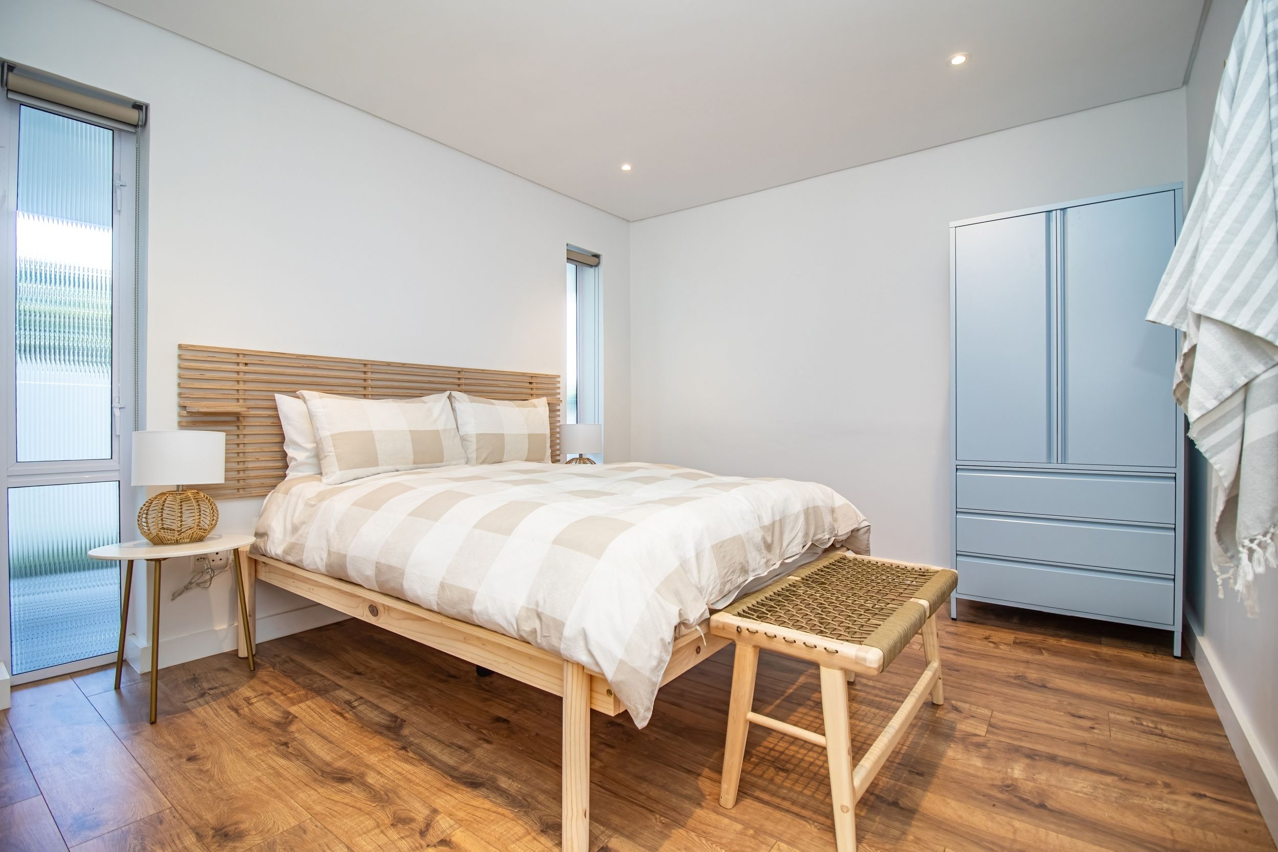 A bedroom with a wooden bed, bedside tables, a bench, a wardrobe, and a window with blinds.
