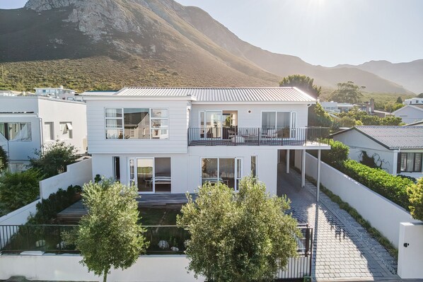 A modern two-story house with a balcony, surrounded by greenery and mountains in the background.