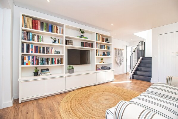 A modern living room with a large bookshelf, a flat-screen TV, and a staircase. - Legacy Villa | Voelklip | Sea & mountain views (Hermanus)