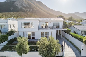 Una casa moderna de dos pisos con balcón, rodeada de vegetación y montañas al fondo.