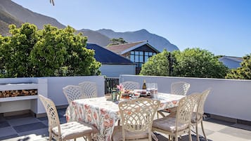 A rooftop terrace with a table set for dining, surrounded by white chairs, and a view of mountains in the background.