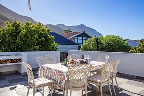 A rooftop terrace with a table set for dining, surrounded by white chairs, and a view of mountains in the background. - Legacy Villa | Voelklip | Sea & mountain views (Hermanus)