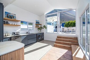 A modern kitchen with built-in appliances, wooden shelves, and large windows offering a view of the outdoors. - Legacy House | Sea and mountain views | Voelklip (Hermanus)