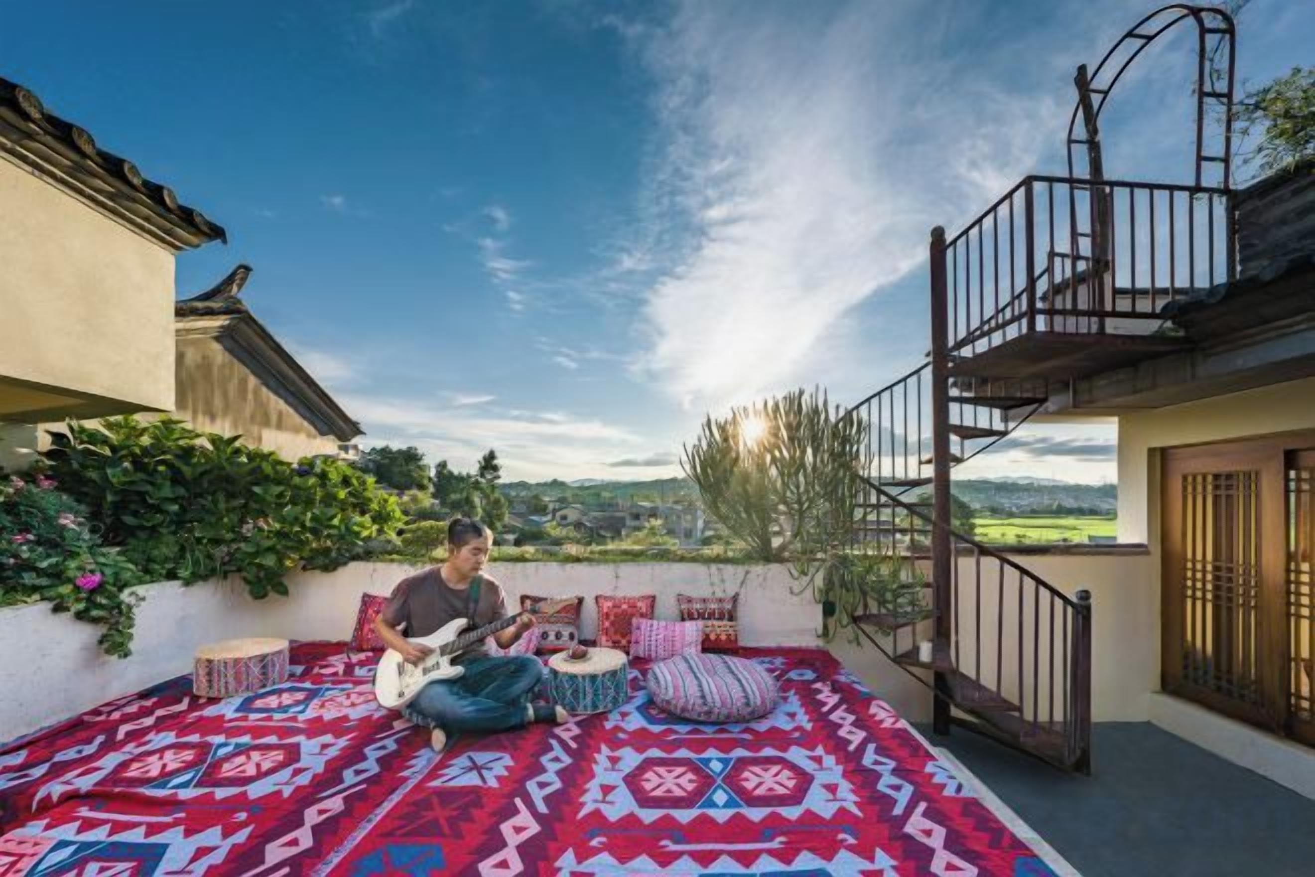 Une personne assise sur un tapis d’extérieur à motifs, jouant de la guitare, avec un balcon et une vue sur la campagne.