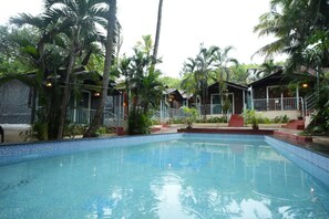 A swimming pool surrounded by palm trees and wooden cabins. - Ashvem Beach Resort (Ashvem)
