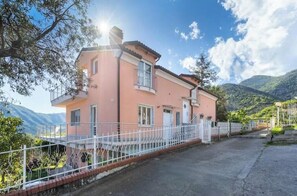 A two-story house with a balcony, surrounded by a white fence, set against a mountainous backdrop. - Hostdomus - Overseas Suite (Boissano)