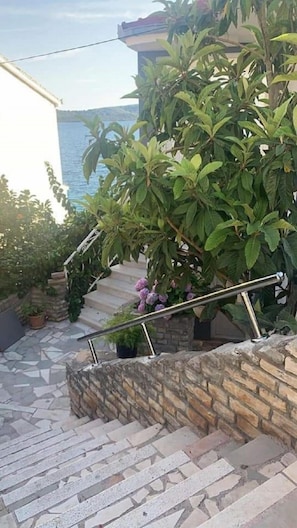 Eine Treppe mit Steinstufen führt zu einem Balkon, umgeben von üppigem Grün und Blick aufs Meer.