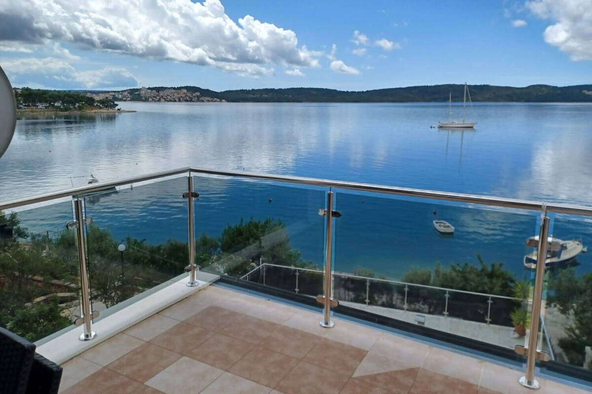 A balcony with a glass railing overlooking a calm lake, a distant shoreline, and a clear sky.