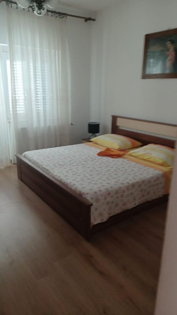 A bedroom with a wooden bed, white curtains, a nightstand, and a painting on the wall. - Apartments Marija (Trogir)