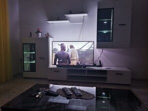 A modern living room with a flat-screen TV on a white entertainment unit, a glass coffee table with remote controls, and a small cabinet with glass doors. - Apartments Marija (Trogir)