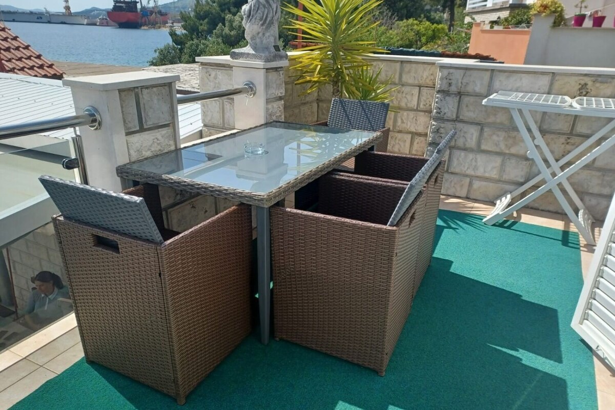 A balcony with a glass table and wicker chairs, overlooking a body of water and a cityscape.