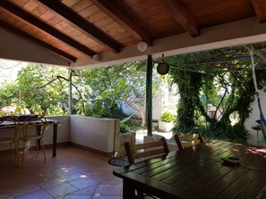 Eine Terrasse im Freien mit einem Holztisch und -stühlen, umgeben von Grünpflanzen und Blick auf Bäume.
