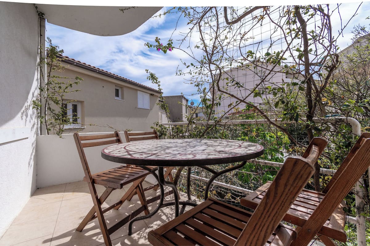 A patio with a round table and wooden chairs, surrounded by a white wall and a garden with a tree and a fence.