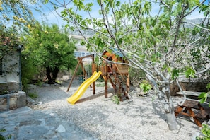Ein Spielplatz mit Rutsche und Schaukeln, umgeben von Bäumen und einem Steinweg.