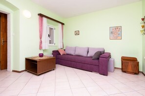 Ein Zimmer mit einem violetten Sofa, einem Holzschrank, einem kleinen Tischchen mit einer violetten Schale, einem Fenster mit Vorhängen und einem Wandbild.