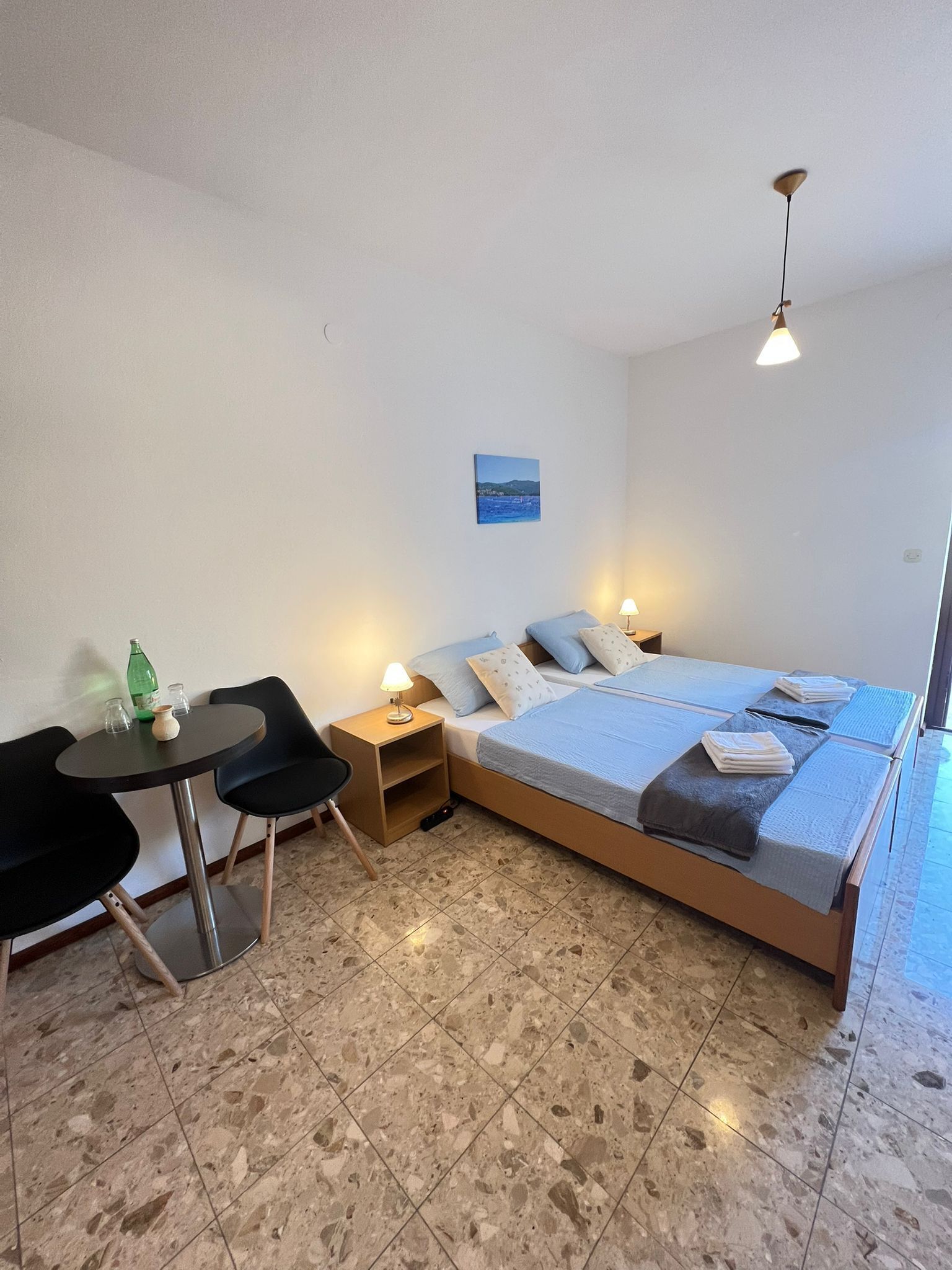 Ein Schlafzimmer mit einem Bett, Nachttischen, einem kleinen Tisch mit Stühlen, einem Bild an der Wand und einem gefliesten Boden.