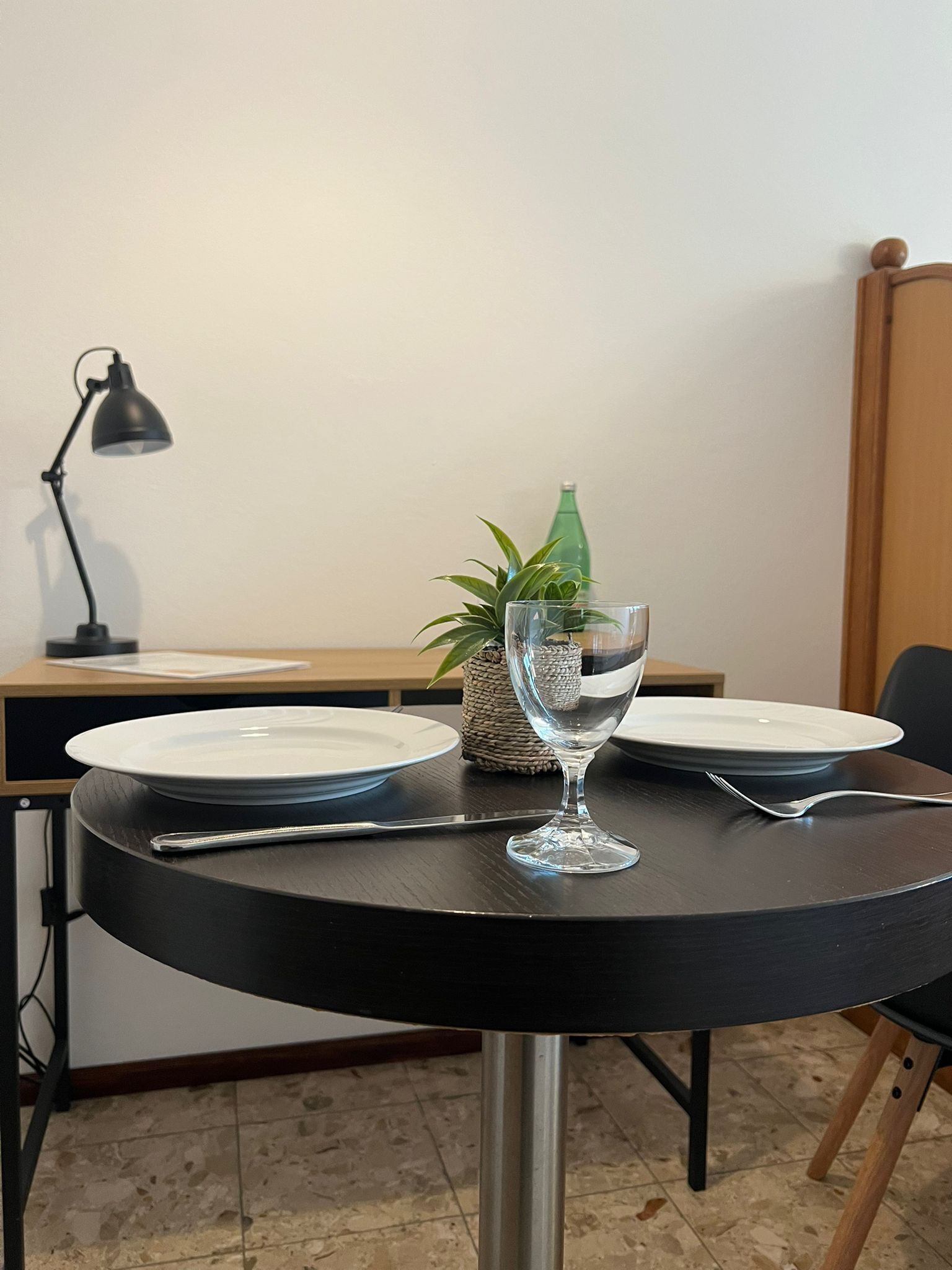 Ein Esstisch mit Tellern, einem Glas, Besteck, einer Lampe und einer kleinen Pflanze.
