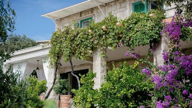 Ein Steinhaus mit überdachter Veranda, grünen Fensterläden und üppiger Bepflanzung, darunter lila Blumen.