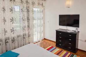 Ein Schlafzimmer mit einem Bett, einer schwarzen Kommode, einem Fernseher und einem Fenster mit durchsichtigen Vorhängen.
