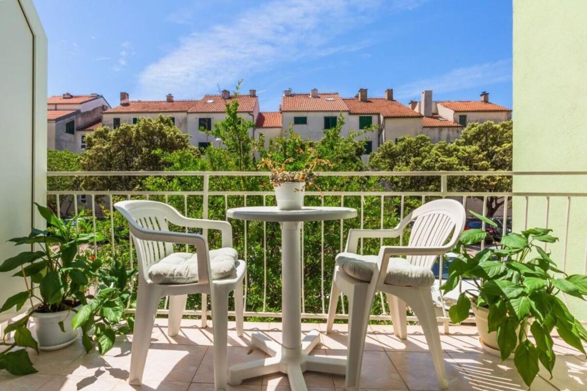 A balcony with two white plastic chairs and a small round table, surrounded by potted plants.
