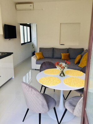 A modern living room with a grey sofa, yellow cushions, a white dining table with a vase of flowers, and a kitchen area with a black TV and a window.