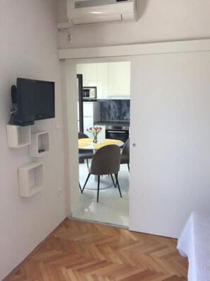 A small room with a flat-screen TV mounted on the wall, a wooden herringbone floor, and a sliding door leading to a kitchen area. - Apartments and Room Tattoo (Makarska)