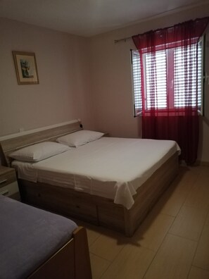A bedroom with a bed, a nightstand, a window with red curtains, and a wall-mounted picture. - Apartment Pavin (Preko)