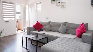 A modern living room with a grey sectional sofa, a small black table, and a red pillow. There are white walls, a window with blinds, and a door leading to another room. - Apartment Sunset (Podstrana)
