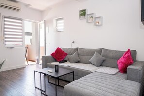 Ein modernes Wohnzimmer mit einer grauen Eckcouch, einem kleinen schwarzen Tisch und einem roten Kissen. Weiße Wände, ein Fenster mit Jalousien und eine Tür zu einem anderen Raum.