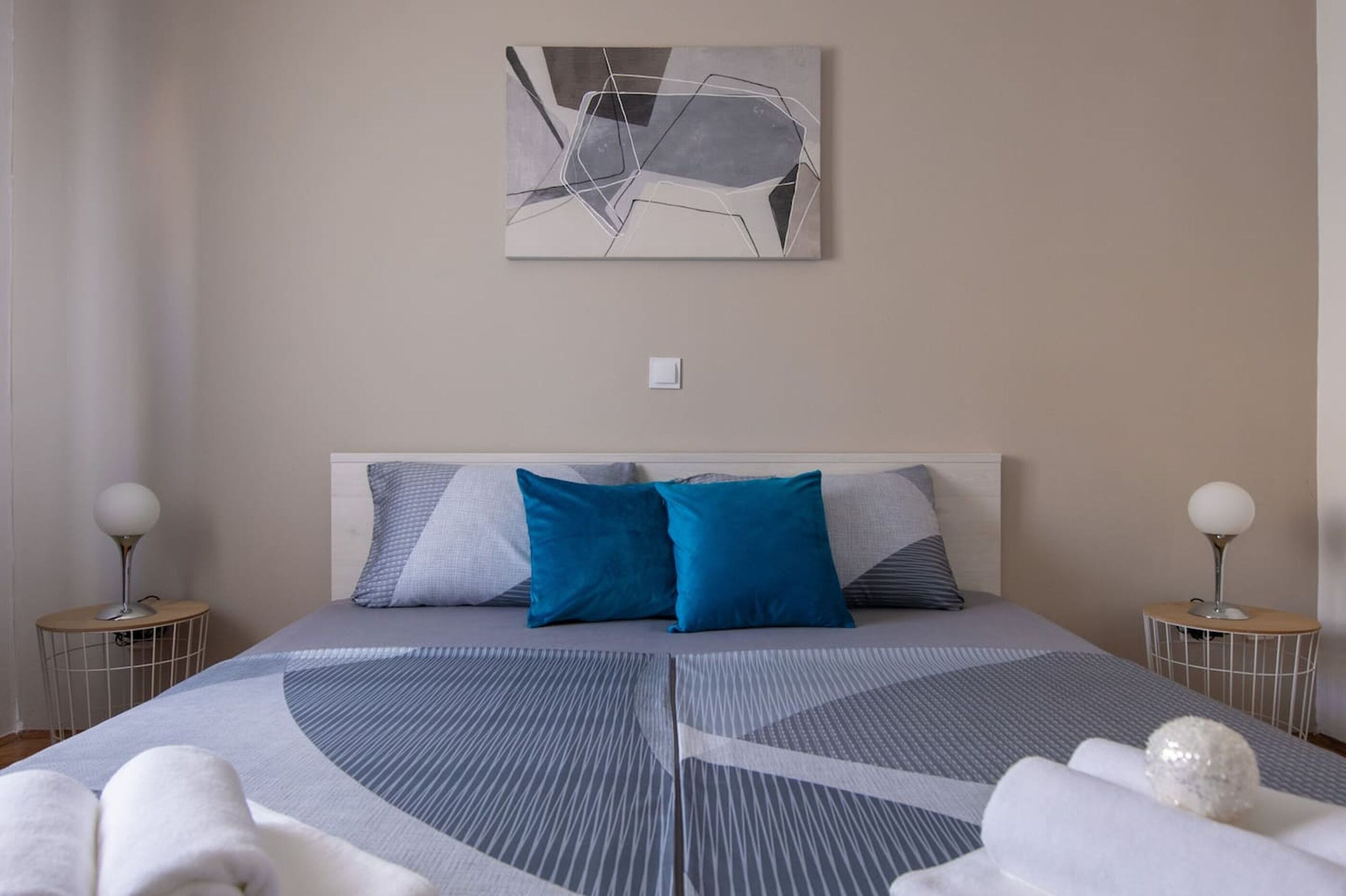 A modern bedroom with a large bed, two bedside tables with lamps, a wall-mounted abstract painting, and a white decorative sphere on the bed.