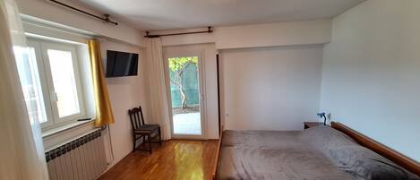 Un dormitorio con suelo de madera, una cama, una silla, un radiador, un televisor y una puerta que da a un balcón.