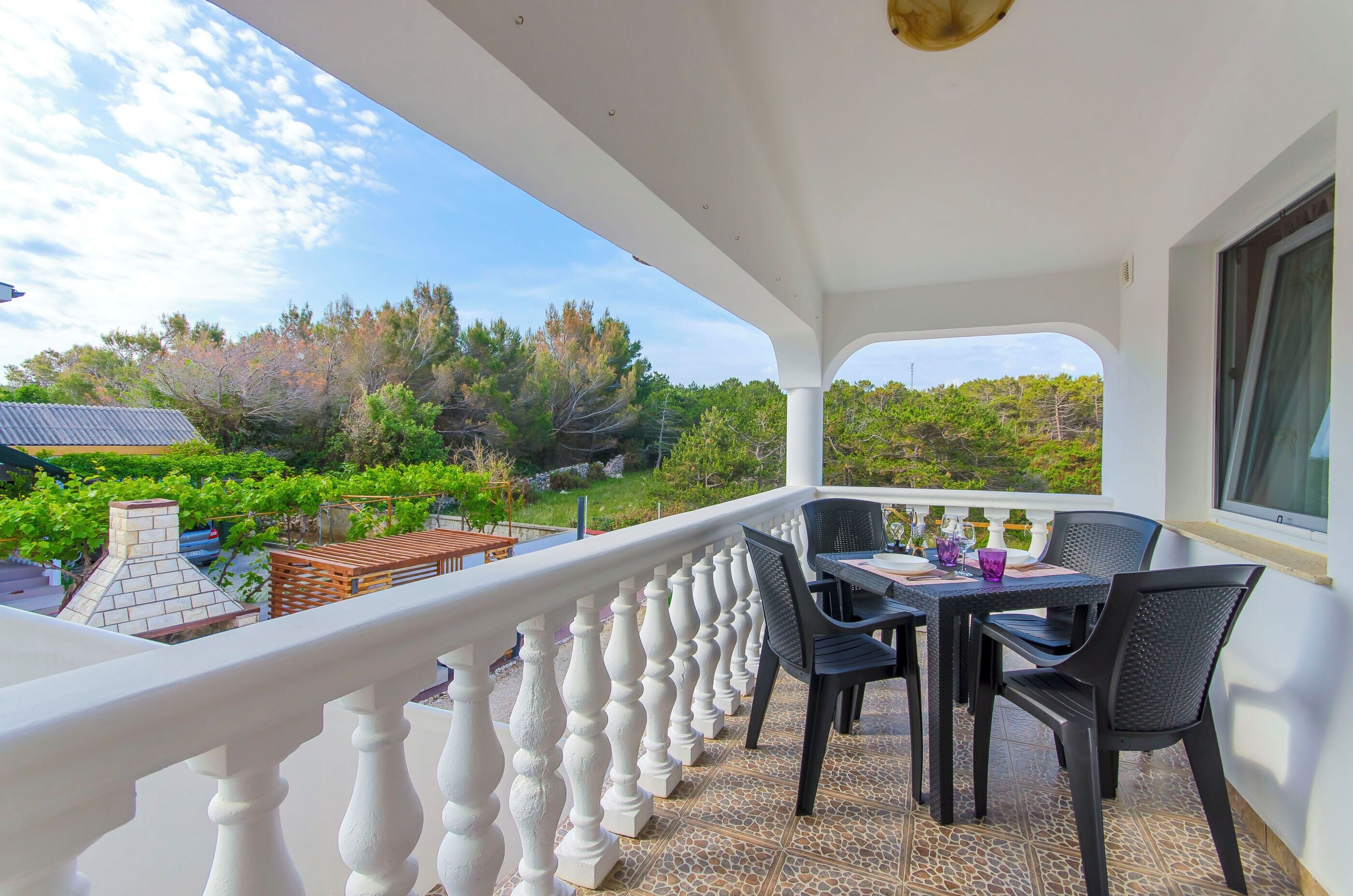 Ein Balkon mit weißem Geländer, einem Tisch mit Stühlen und Blick auf einen Garten und Bäume.