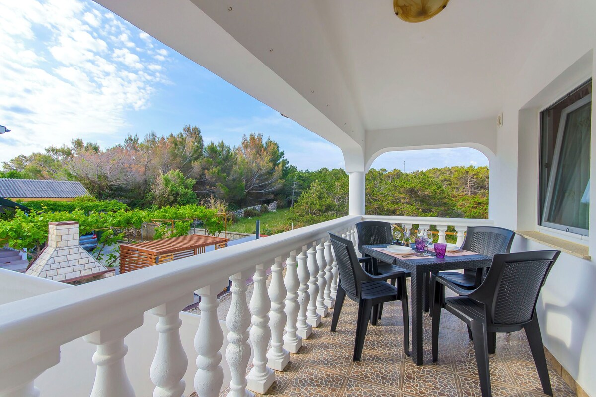 A balcony with a white balustrade, a table with chairs, and a view of a garden and trees.