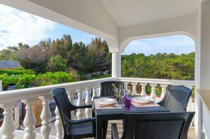A covered outdoor dining area with a table set for two, white balustrades, and a view of trees and a building in the background. - Apartments Apartmani Didovi Dvori (Razanac)