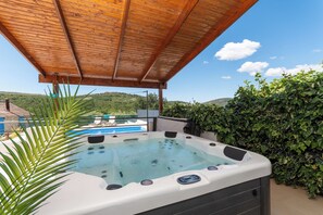 Ein Whirlpool unter einer Holzpergola mit Pool und Grünpflanzen im Hintergrund.