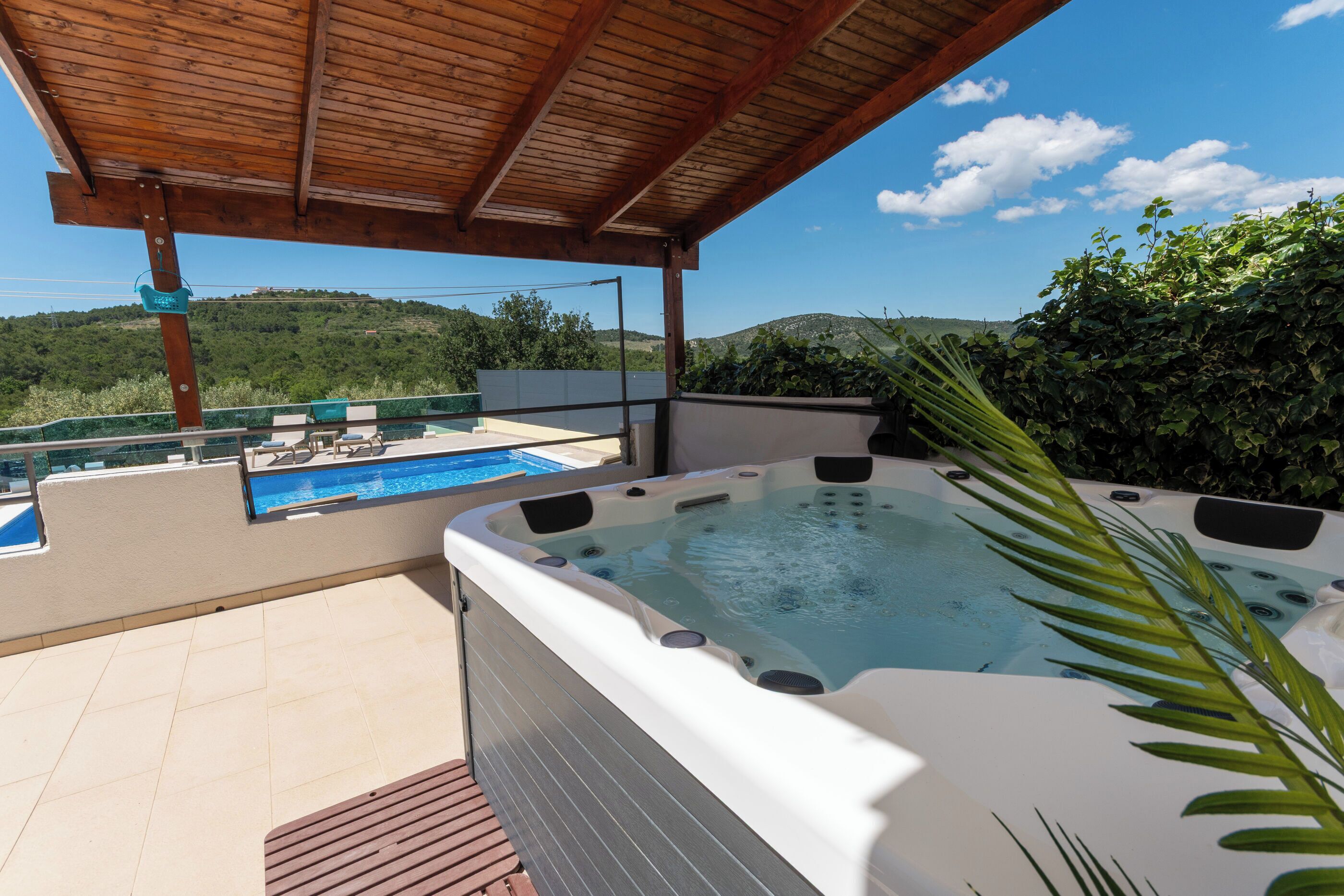 A hot tub, a swimming pool, and a view of hills under a wooden pergola.