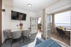 Ein modernes Zimmer mit Flachbildfernseher an der Wand, einem Esstisch mit vier Stühlen, einer Couch und einem Balkon mit Tisch für zwei Personen.