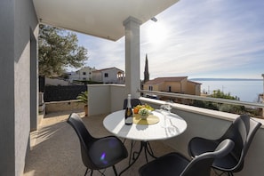 Ein Balkon mit rundem Tisch, Stühlen, einer Flasche und einem Glas, mit Blick auf die Küste, Häuser und einen Kirchturm.