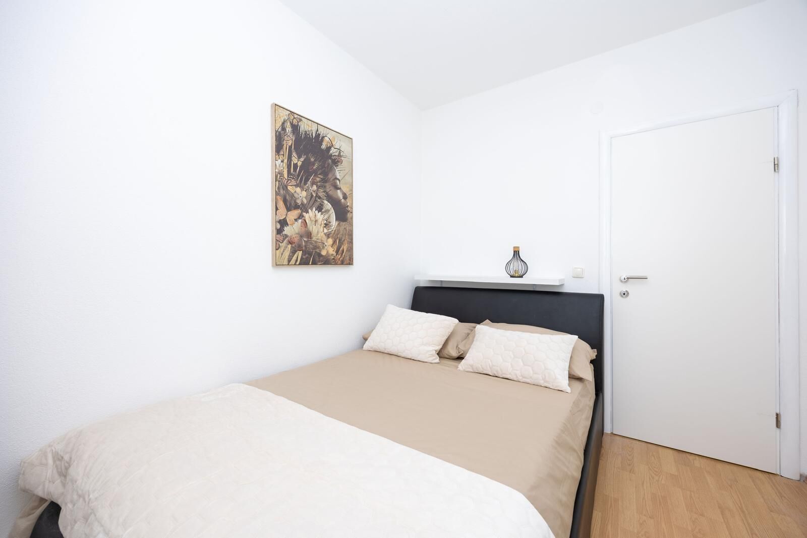 A bedroom with a bed, a door, and a wall-mounted shelf with a vase.
